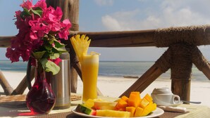 Un tavolo con un piatto di frutta, un bicchiere di succo, un vaso con fiori e una tazza e un piattino, in un contesto balneare.