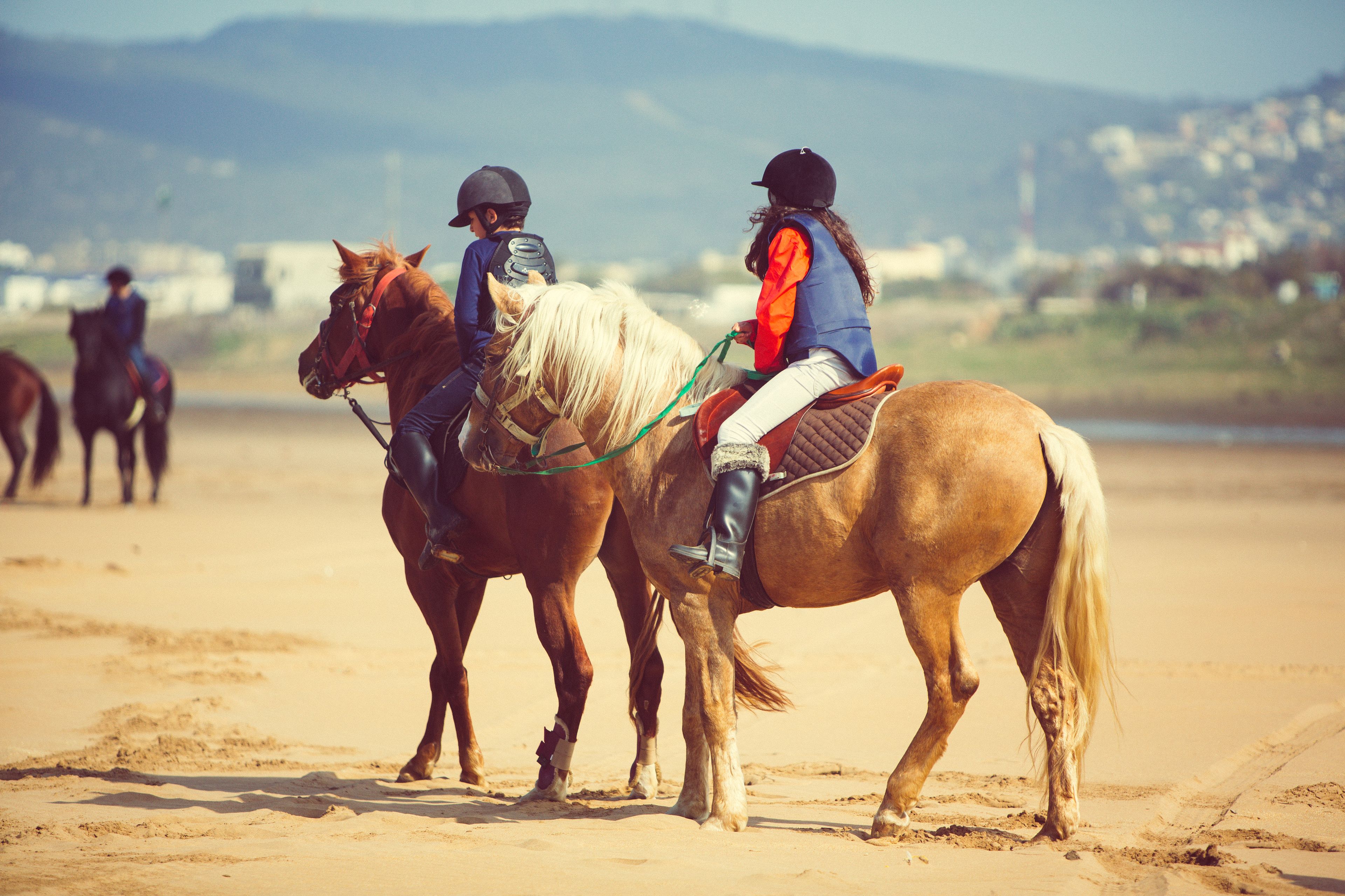 Two people riding horses on a sandy beach with a cityscape in the background.