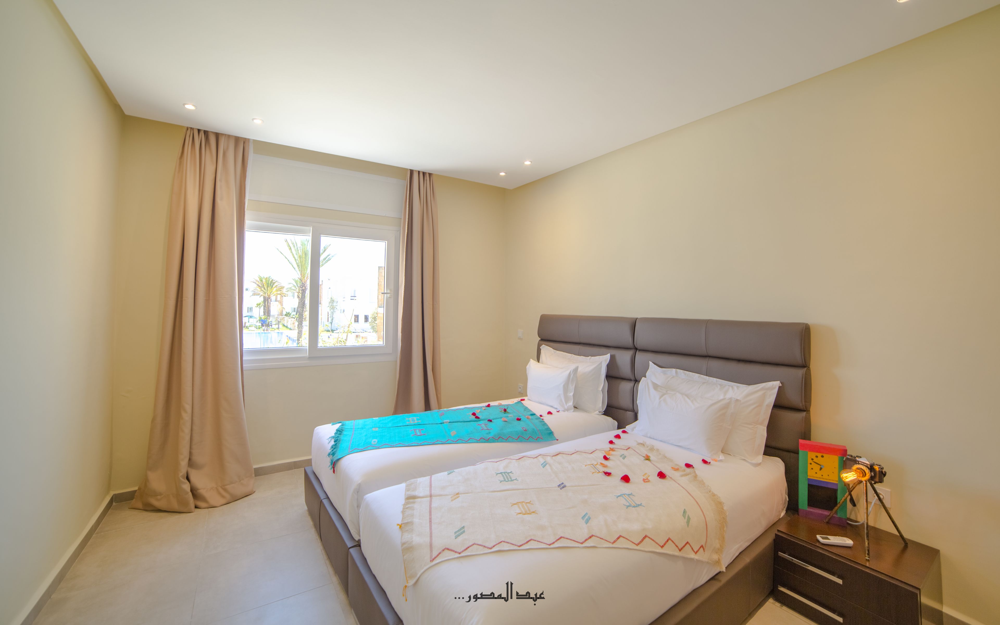Un dormitorio con dos camas, cortinas color beige, una ventana con vistas y una mesita de noche con lámpara.