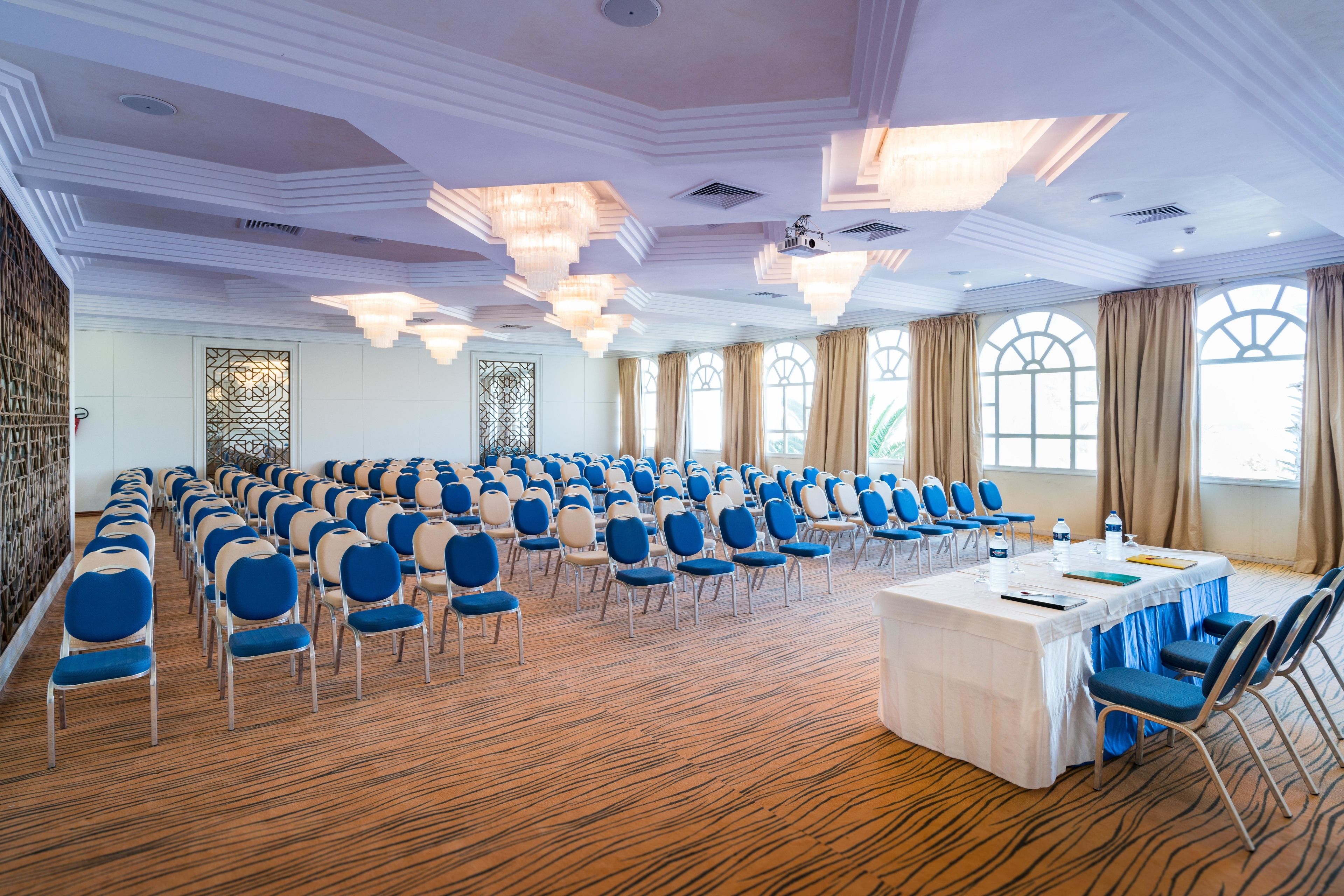 Une salle de conférence avec des rangées de chaises, une table recouverte d’une nappe blanche et de grandes fenêtres avec des rideaux.