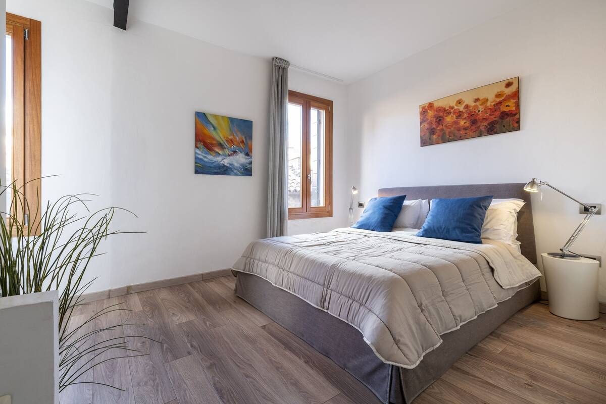 A bedroom with a bed, a window, a painting, and a bedside lamp.