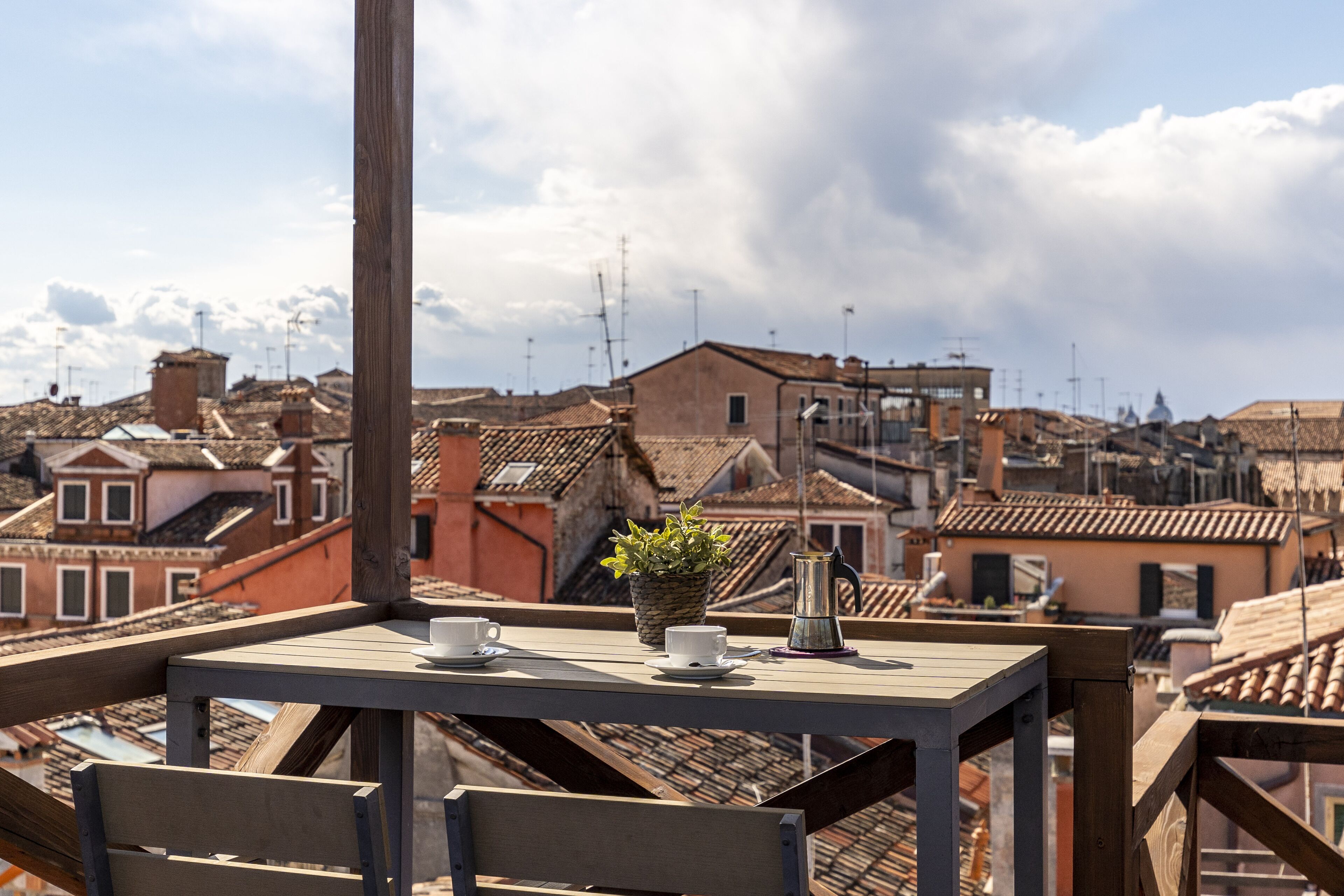 A wooden table with a potted plant, two cups, and a teapot on a balcony overlooking a cityscape with terracotta rooftops.