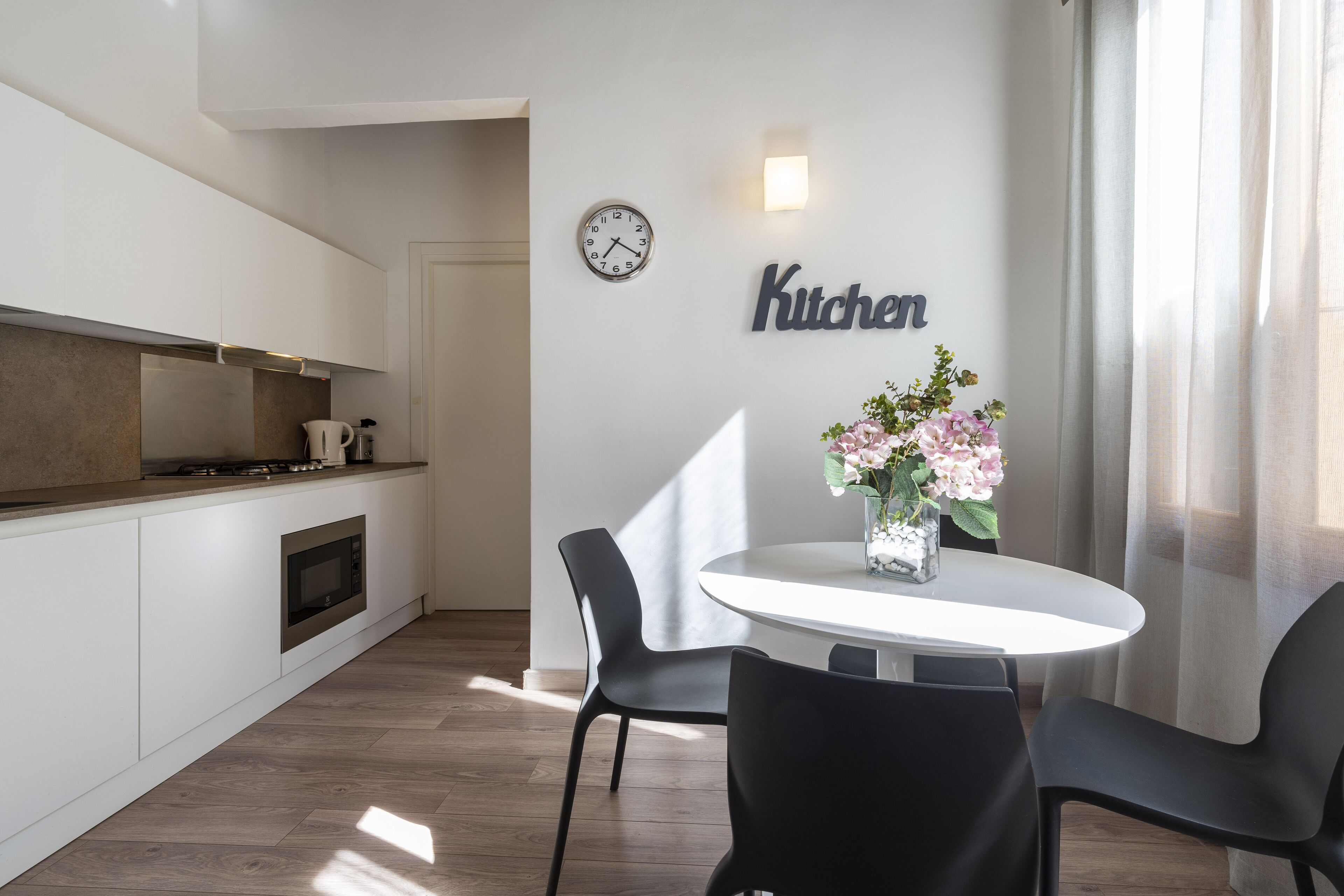 A modern kitchen with a clock, a 'Kitchen' sign, and a dining area with chairs and a table.
