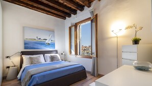 Een slaapkamer met een bed, een schilderij van een zeilboot, een raam met uitzicht, een bedlampje en een klein tafeltje met een kom.