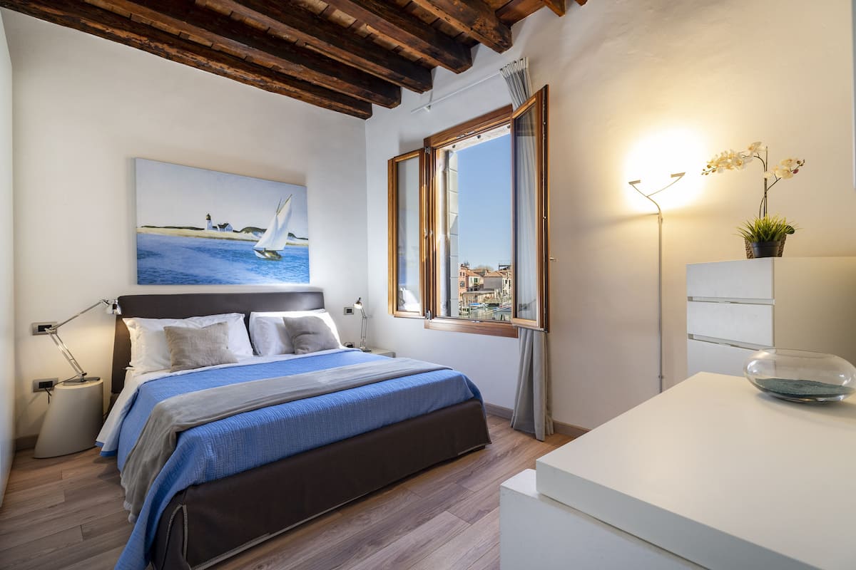 A bedroom with a bed, a painting of a sailboat, a window with a view, a bedside lamp, and a small table with a bowl.