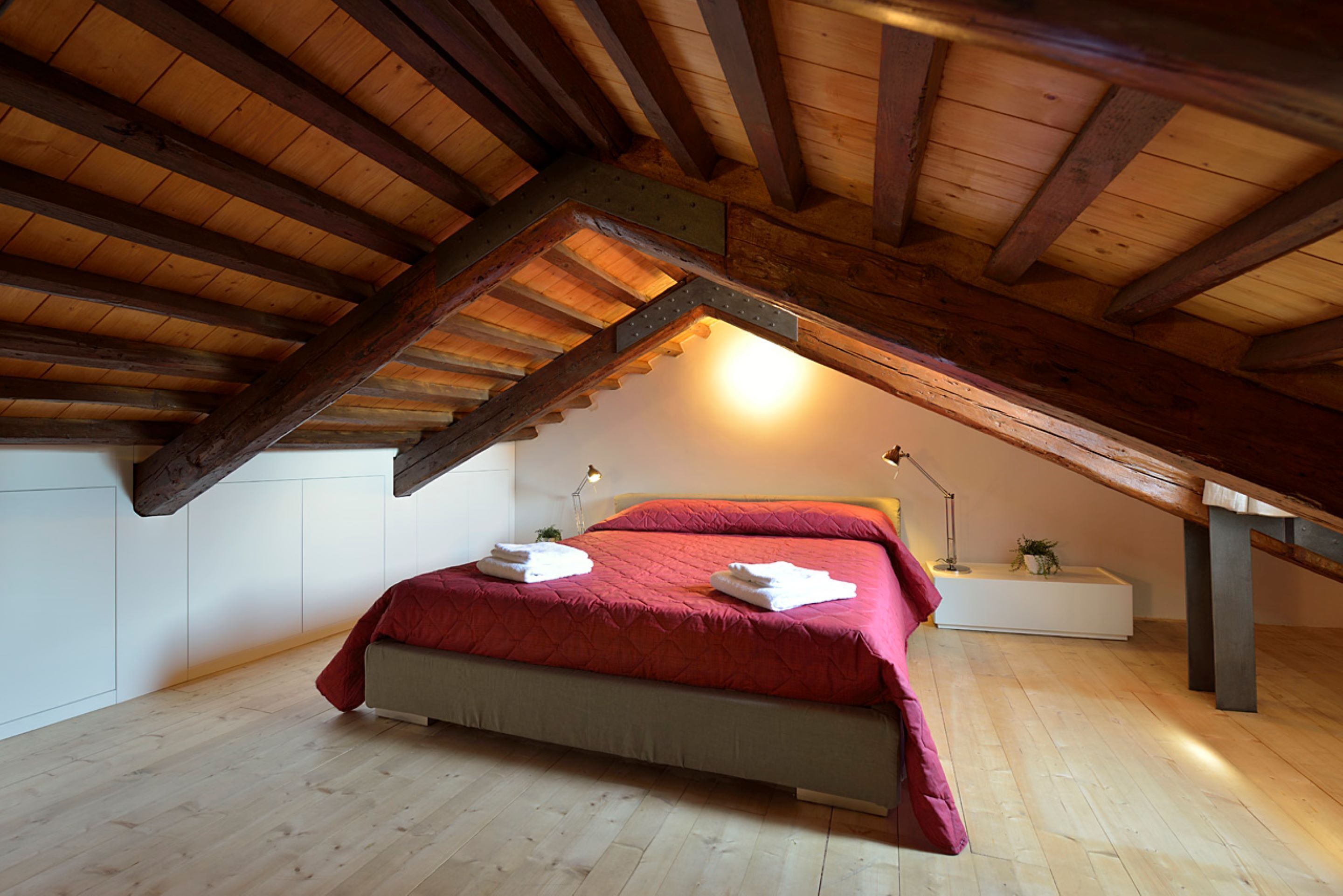 A bedroom with a sloped ceiling, wooden beams, a bed with red bedding, a bedside table, and a lamp.