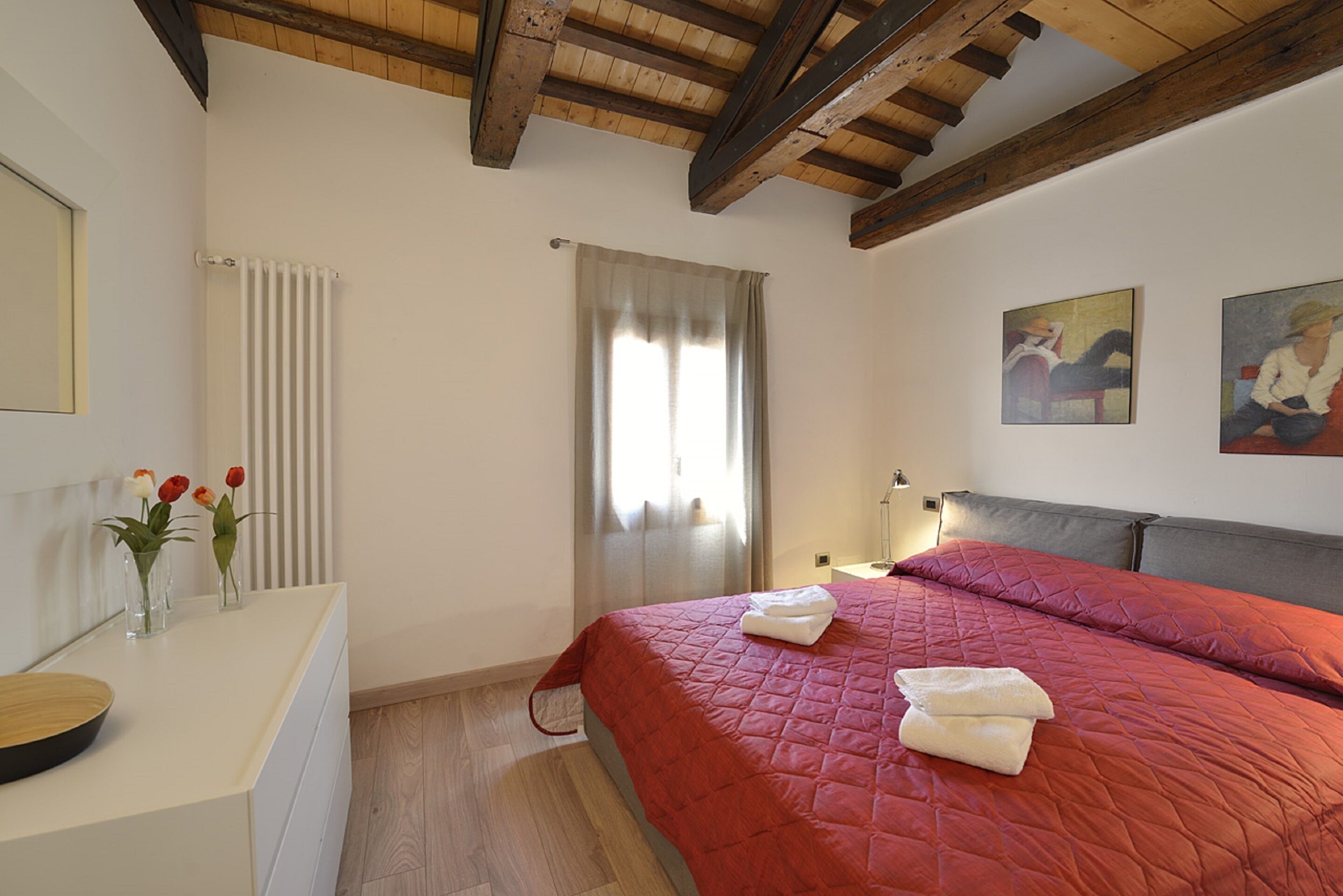 Une chambre avec un plafond en bois, un lit recouvert d’une couette rouge, une table de chevet blanche ornée d’un vase de fleurs et un radiateur.