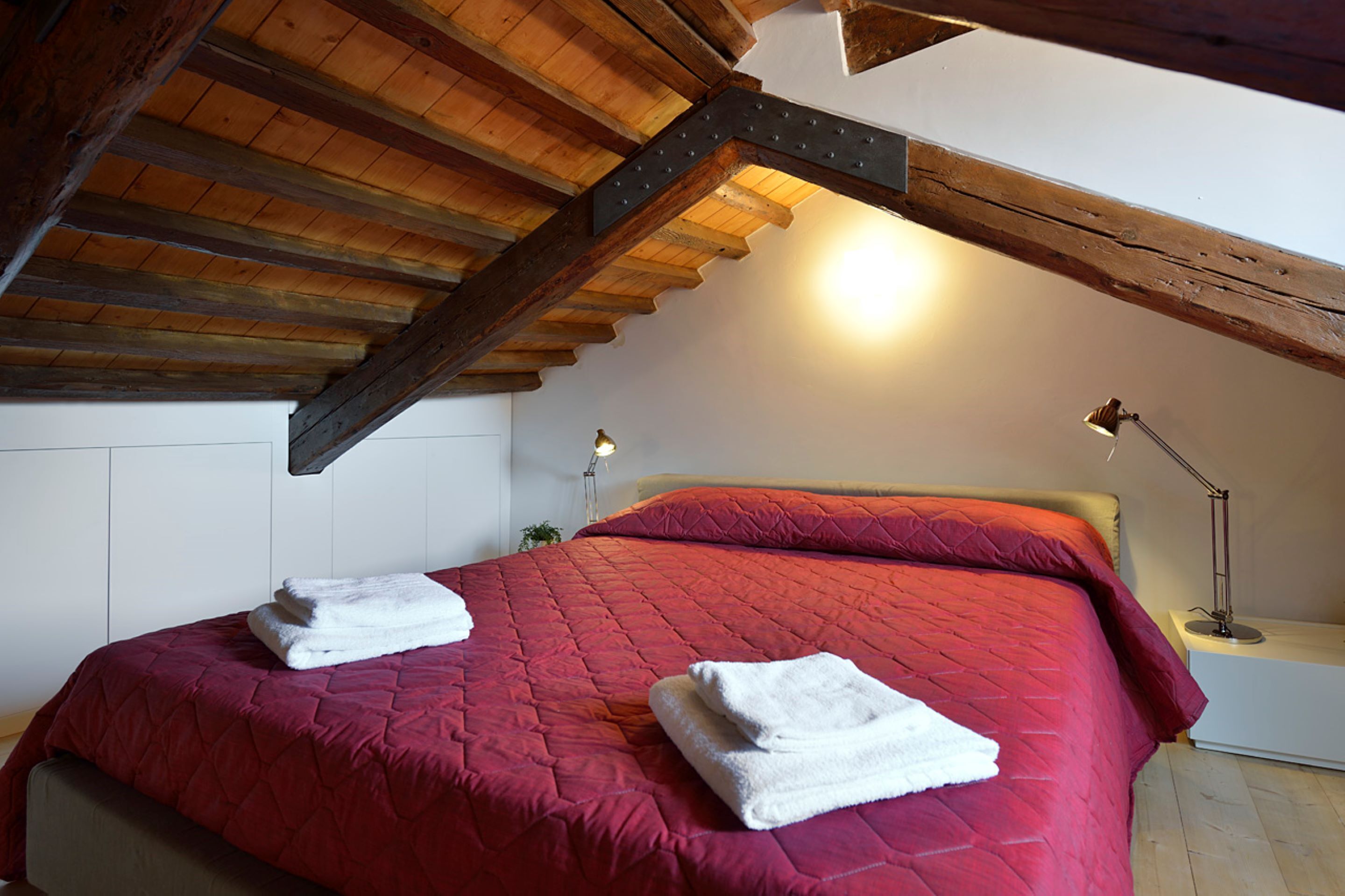 Un dormitorio con techo de madera y una colcha roja. Hay dos toallas blancas sobre la cama.