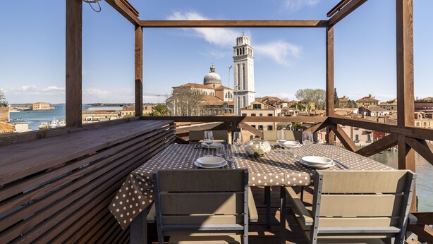 Een houten terras met een tafel gedekt voor twee, met uitzicht op een historisch gebouw met een klokkentoren en een waterpartij.