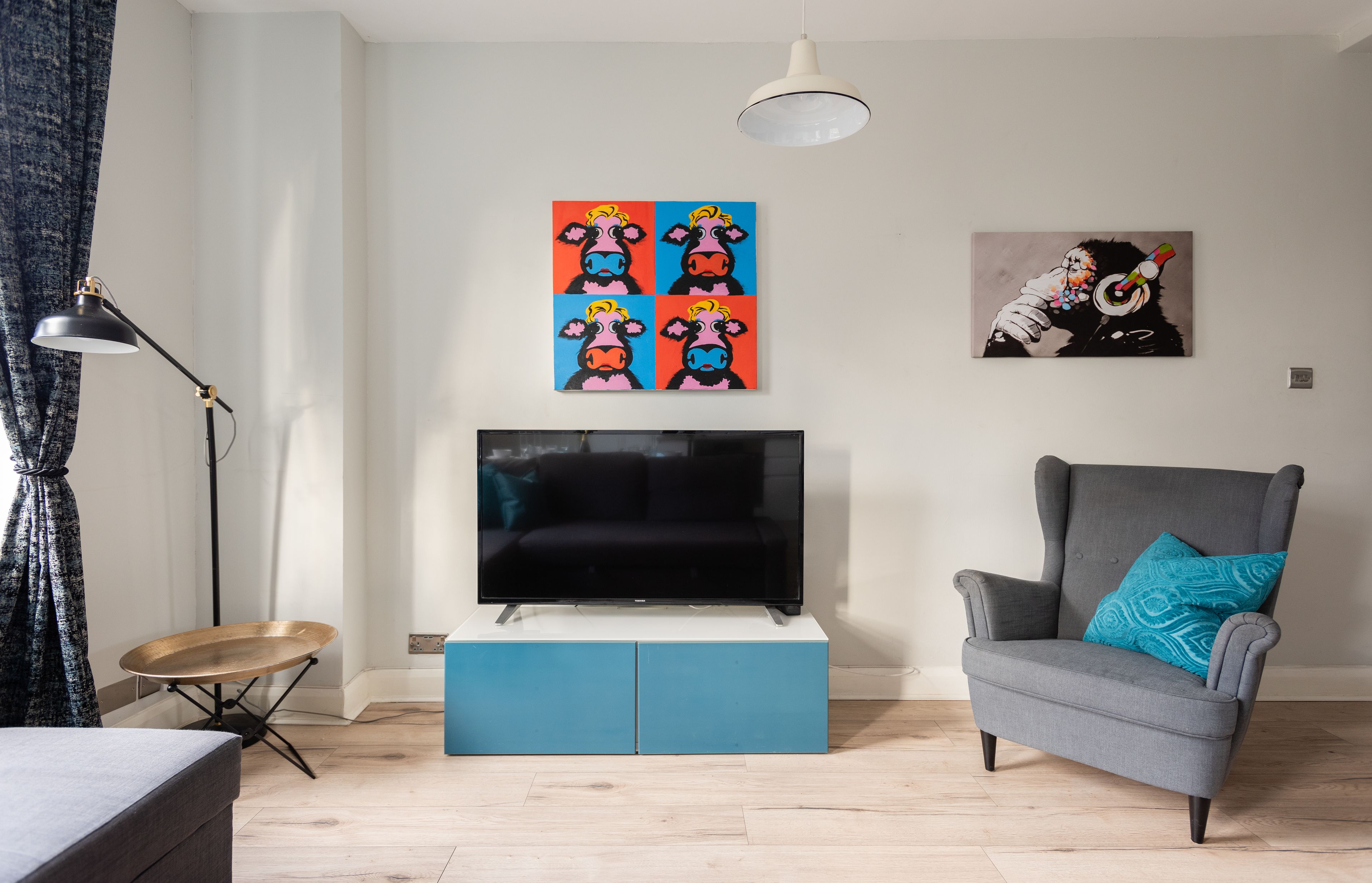 A living room with a flat-screen TV on a blue cabinet, a grey armchair with a blue pillow, a black floor lamp, and two wall art pieces.