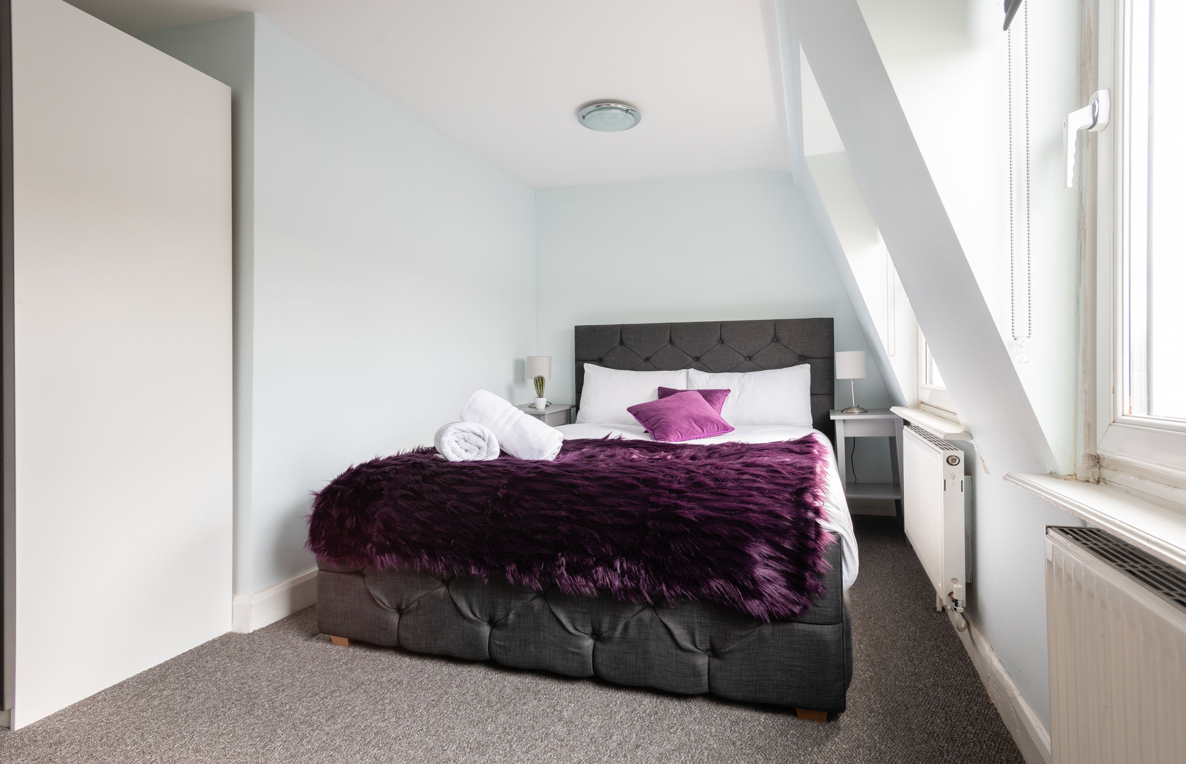 A bedroom with a bed, a nightstand, a lamp, and a window.