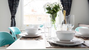 Una mesa de comedor decorada con platos, tazones y cubiertos blancos, un florero con flores blancas y una botella de champán.
