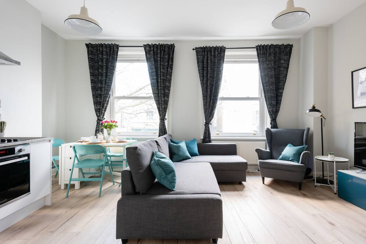 A modern living room with a grey sectional sofa, teal chairs, a dining table, and a kitchen area with a stove and cabinets.