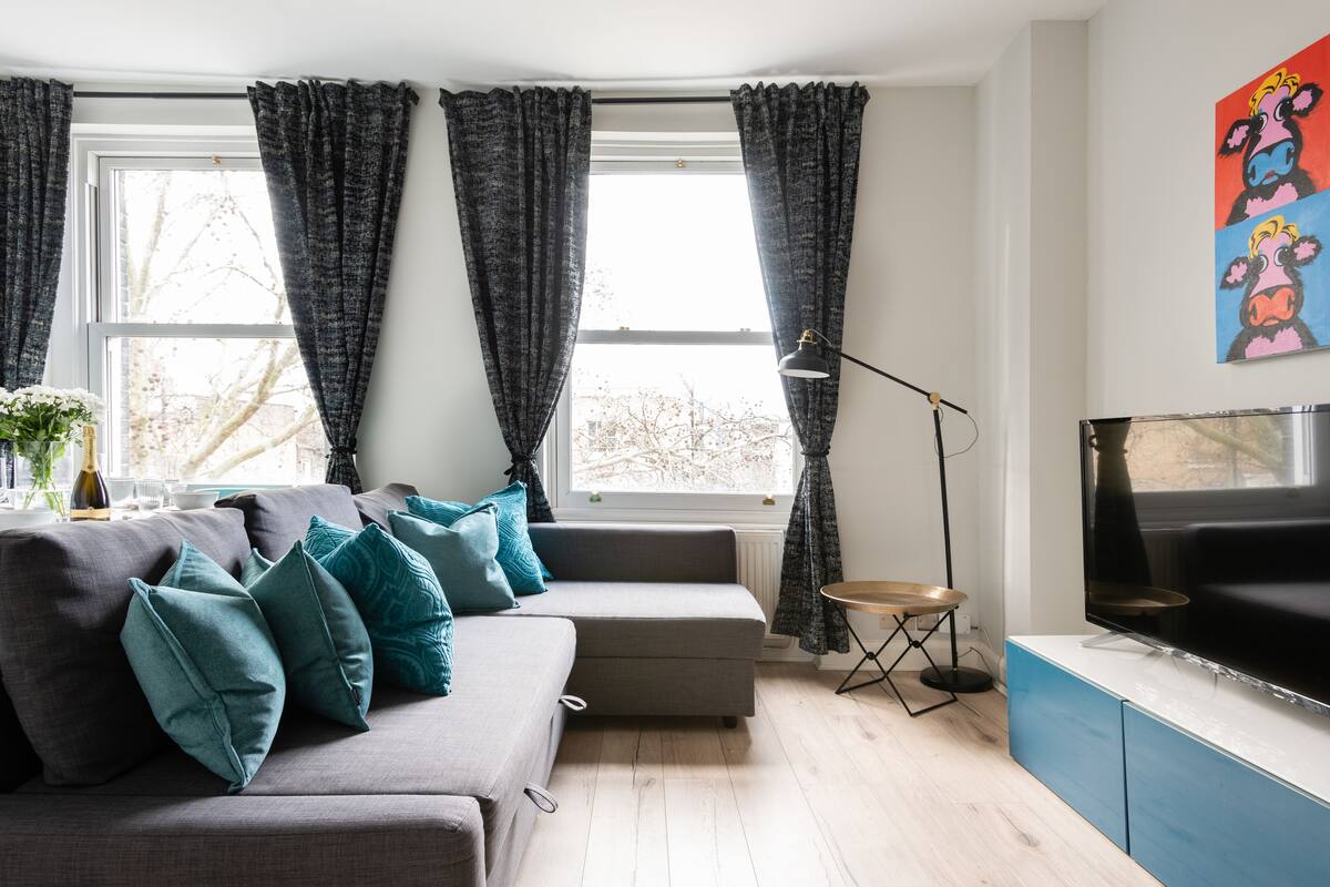 A modern living room with a grey sofa, teal cushions, a small side table, a floor lamp, and a TV on a blue cabinet.