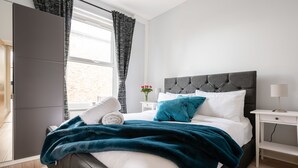 Una cama bien hecha con cabecera gris, ropa de cama blanca y una manta turquesa. Se aprecia una mesita de noche con una lámpara y un jarrón con flores. Se ve una ventana con cortinas, y un armario está parcialmente abierto a la izquierda.