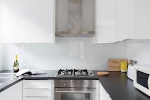 Eine moderne Küche mit weißen Schränken, einem Gasherd, einem Backofen, einer Mikrowelle und einer kleinen Kochinsel mit Korktafel und gelber Aufbewahrungsbox.