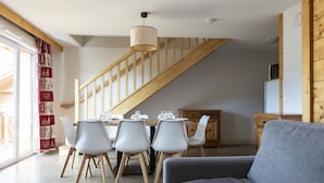 A modern living room with a grey sofa, a dining area with white chairs and a wooden table, and a staircase with wooden railings.
