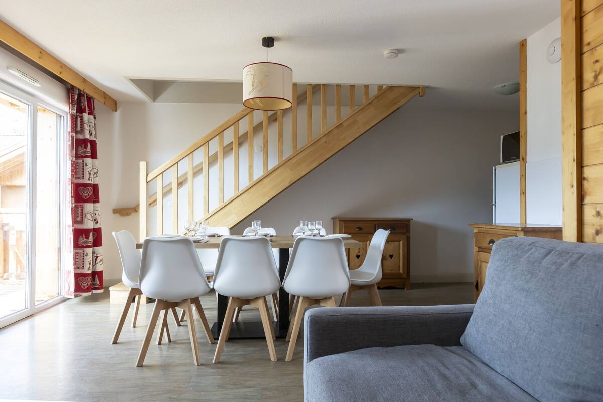 Ein modernes Wohnzimmer mit einem grauen Sofa, einem Essbereich mit weißen Stühlen und einem Holztisch sowie eine Treppe mit Holzbalken.