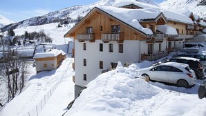 A snow-covered building with multiple balconies, a ski slope in the background, and parked cars.