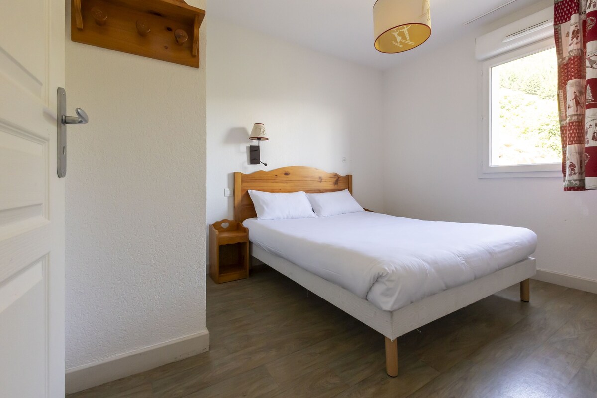 Un dormitorio con una cama de madera, una mesita de noche, una lámpara de pared y un ventanal con cortinas.
