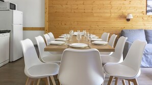 A dining area with a wooden table set for 8, surrounded by white chairs. A kitchen with a refrigerator and stove is visible to the left.