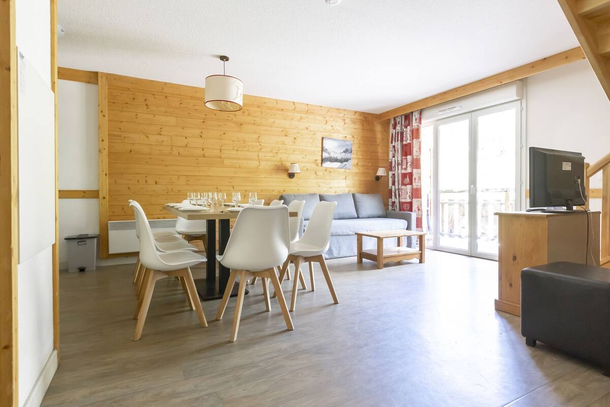 Ein modernes Wohnzimmer mit einer Holzwand, einem Essbereich mit weißen Stühlen, einer Couch und einem Fernseher auf einer Holzkommode.
