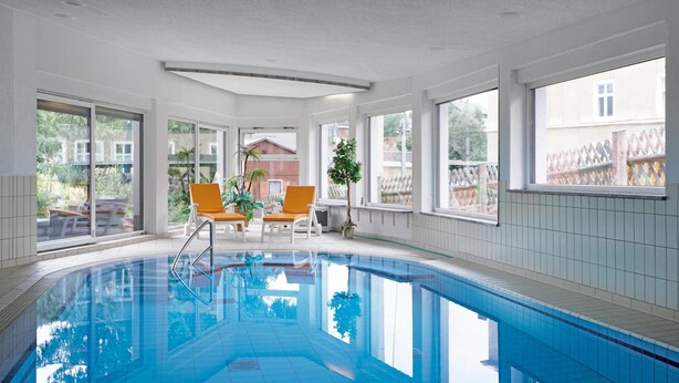 Una piscina interior con paredes de vidrio transparente, dos sillas anaranjadas y una planta en maceta.