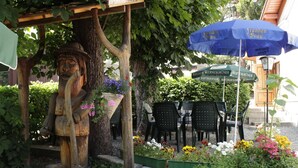 Una estatua de madera bajo una pérgola, zona de estar al aire libre con mesas y sillas, y un paraguas azul.