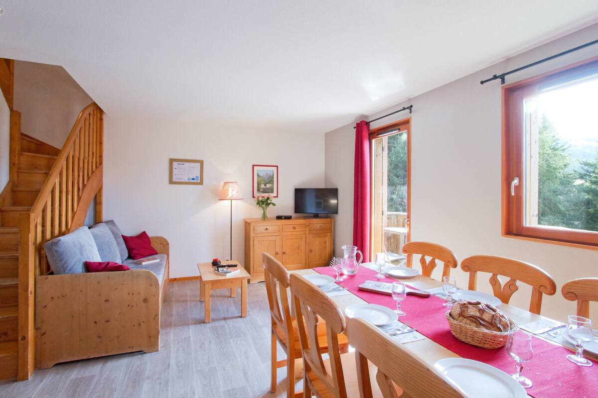 Ein gemütliches Zimmer mit einer Holztreppe, einer Couch mit Kissen, einem Esstisch für zwei Personen und einem Fernseher auf einer Holzkommode.