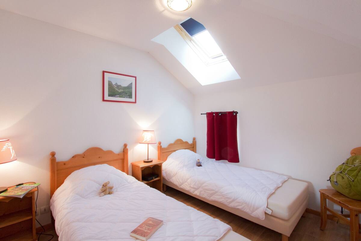 Ein Schlafzimmer mit zwei Einzelbetten, einem Oberlicht, einem Nachttisch mit Lampe, einem gerahmten Bild an der Wand und einer grünen Tasche auf einem kleinen Tisch.