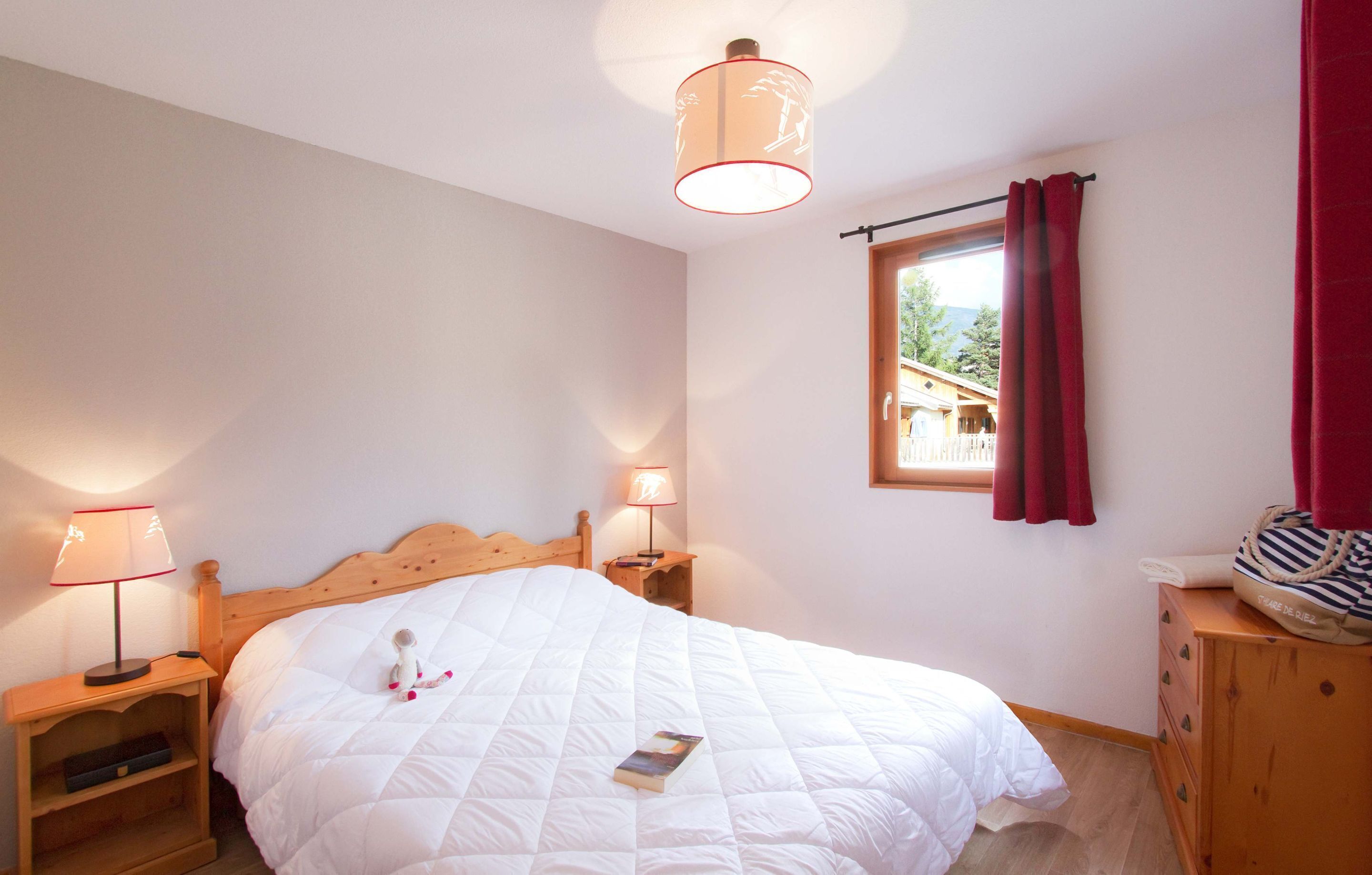 A bedroom with a wooden bed, two bedside lamps, a window with curtains, and a dresser with a striped bag on top.
