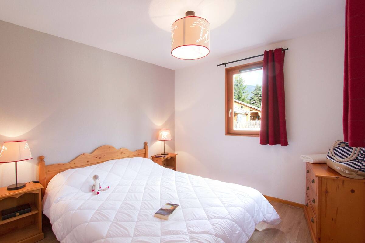 Ein Schlafzimmer mit einem hölzernen Bett, zwei Nachttischlampen, einem Fenster mit Vorhängen und einer Kommode mit einer gestreiften Tasche darauf.