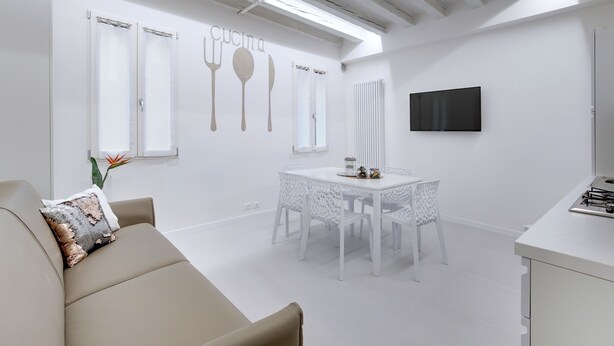 Una cocina moderna con armarios blancos, una mesa de comedor y sillas. Hay un televisor montado en la pared y una ventana con persianas.