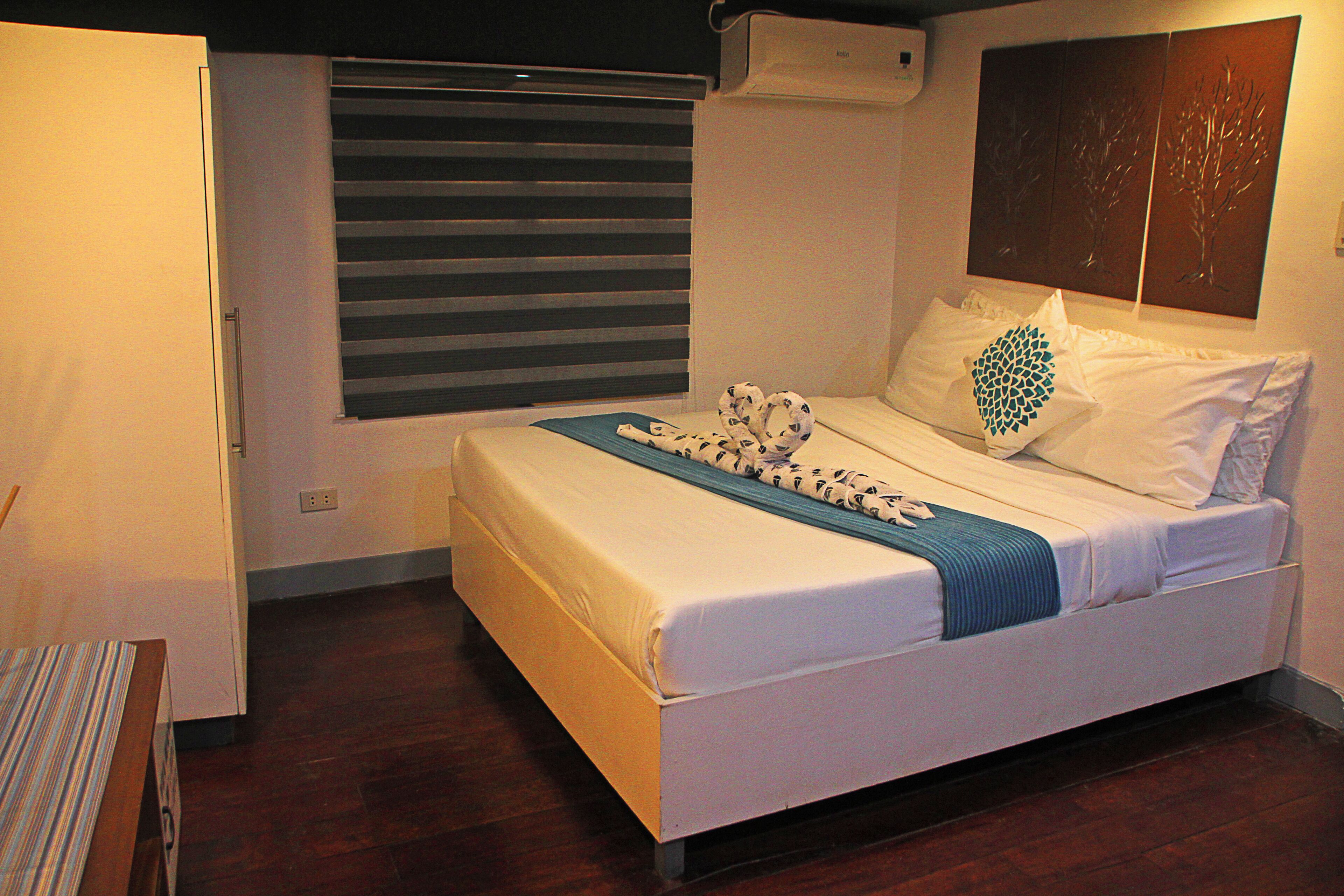 Un dormitorio con cama, ventana con persianas, aire acondicionado de pared y una mesita con mantel a rayas.