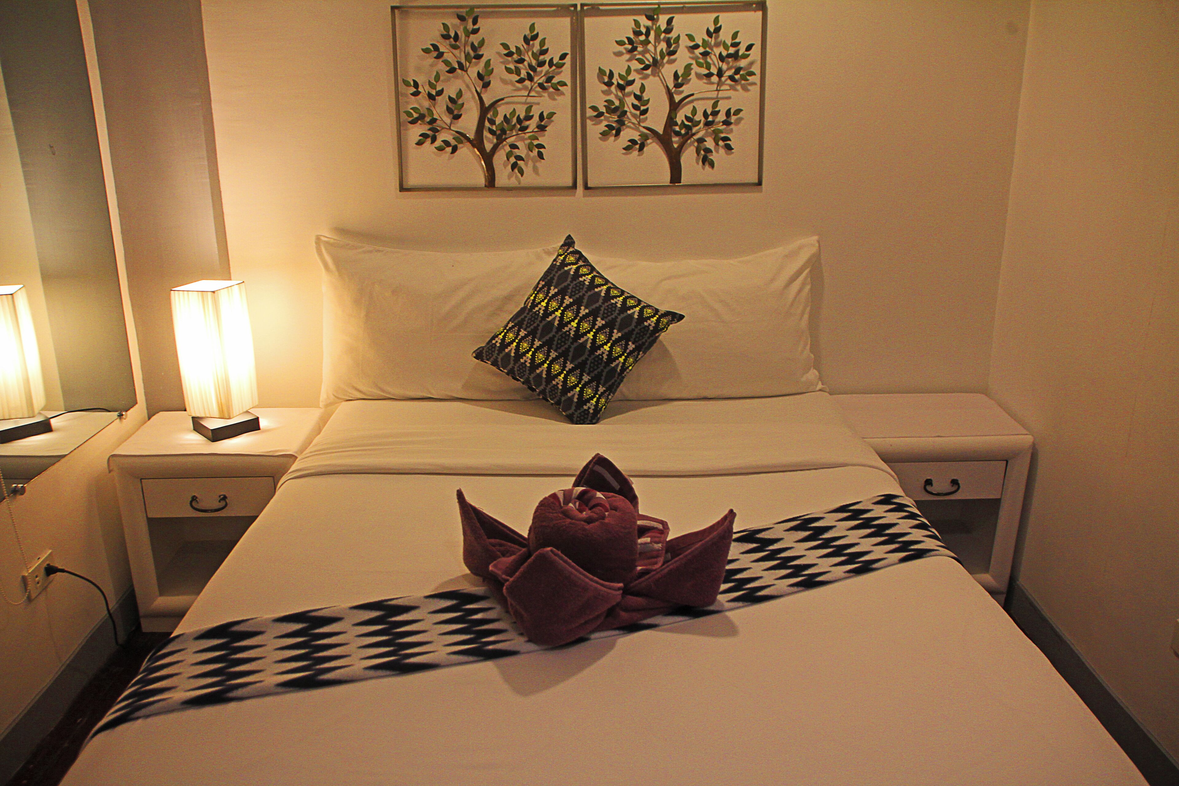 A neatly made bed with a patterned bedspread, a decorative pillow, and two wall-mounted paintings of trees.