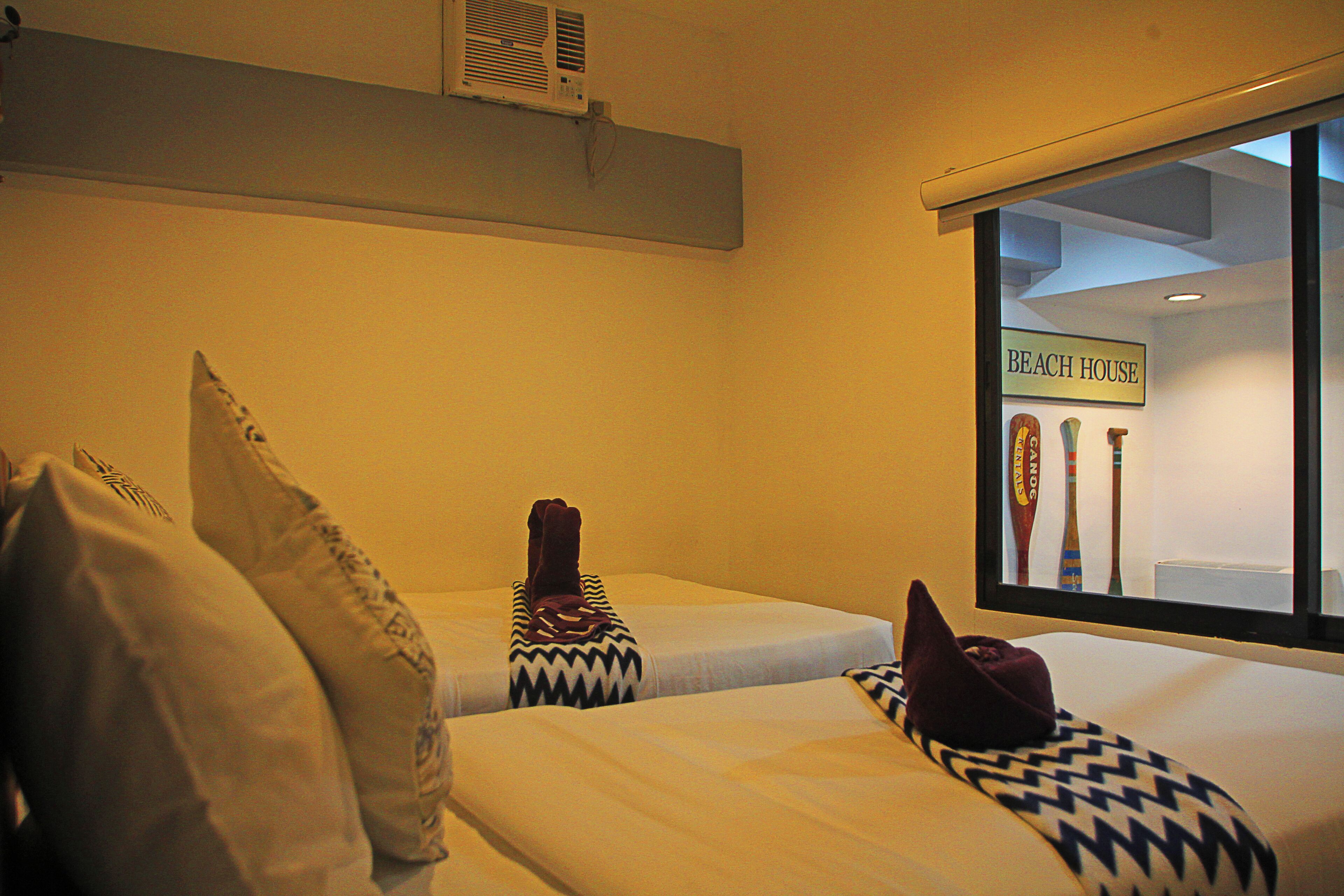 A hotel room with two beds, each with patterned pillows, a window with a view of the beach, and a sign reading "BEACH HOUSE".