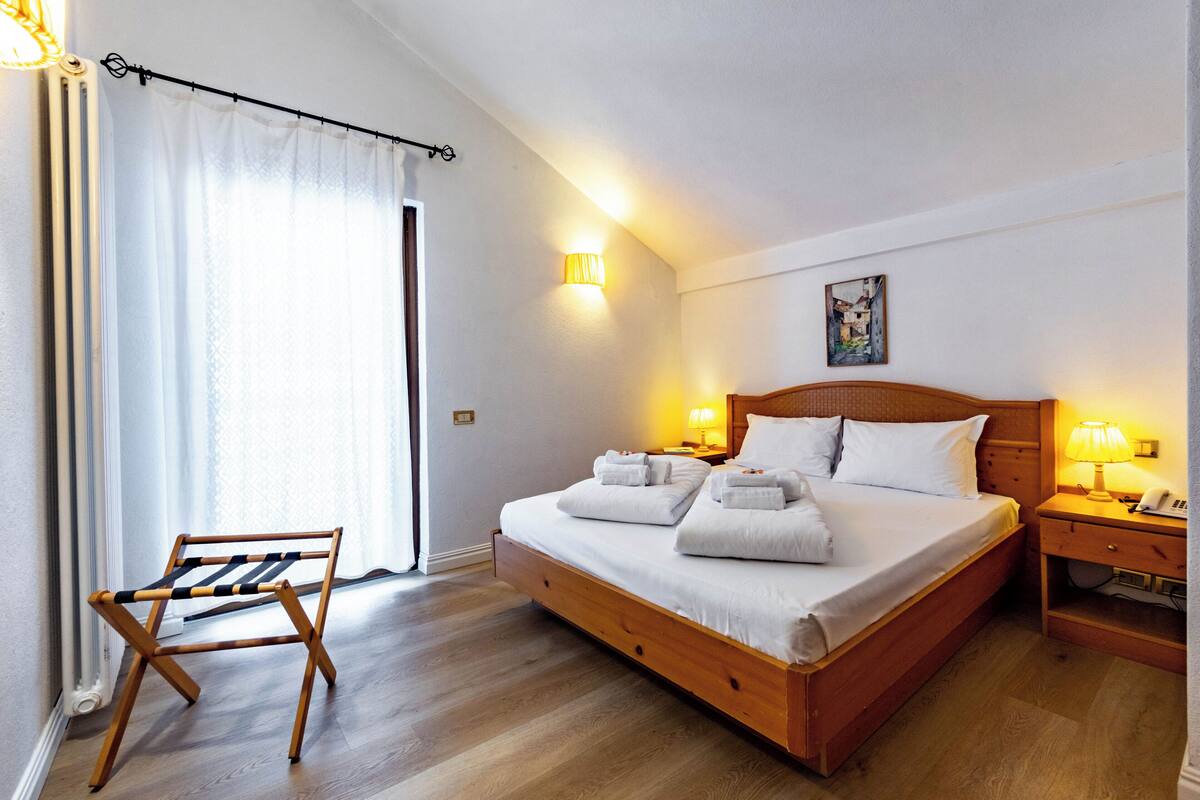 A neatly made bed with white linens and pillows, a wooden nightstand with a lamp, and a wooden chair with a striped seat.