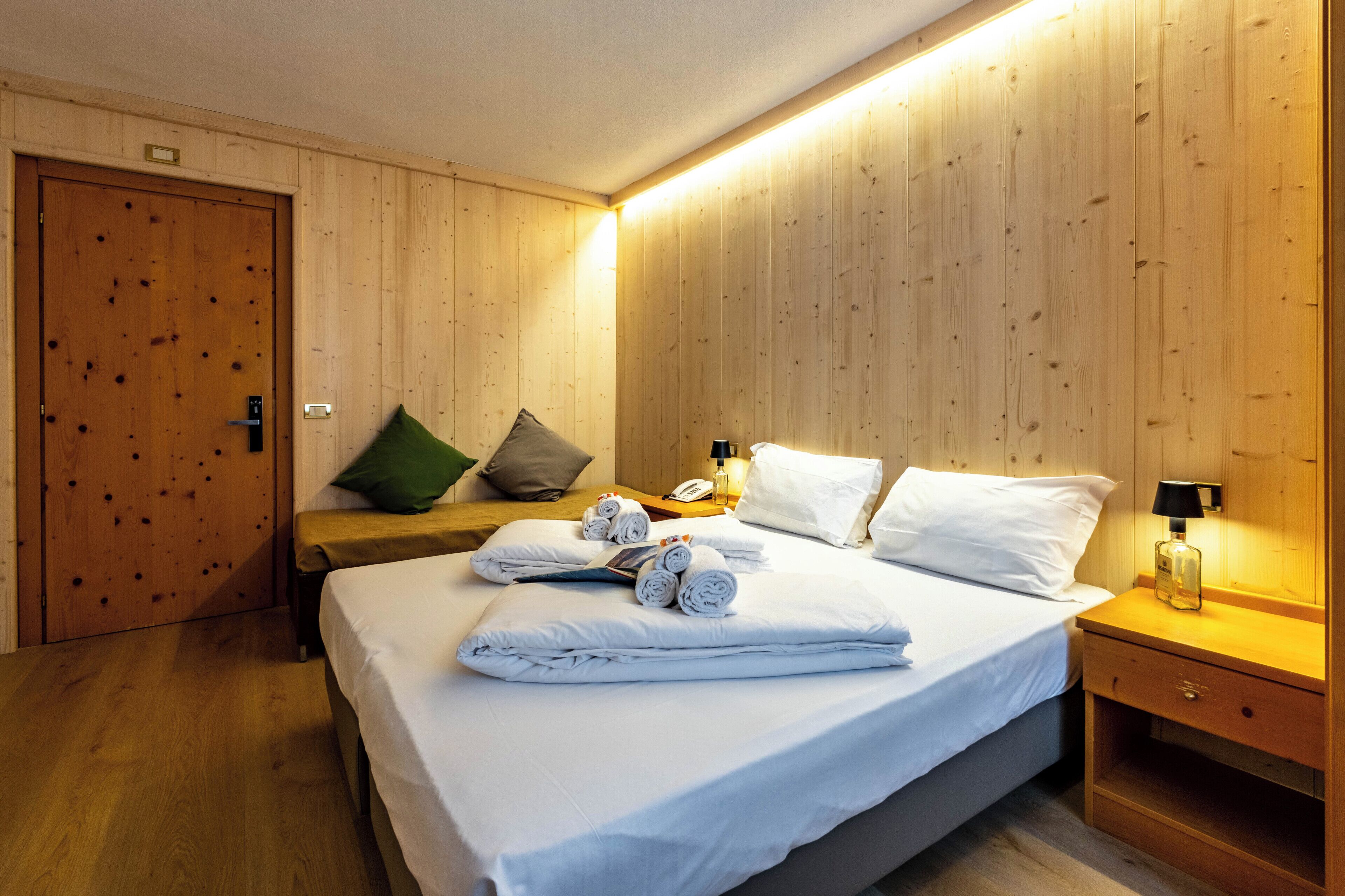 A hotel room with a wooden panel wall, a bed with white linens, a bedside table with a lamp, and a wooden door.