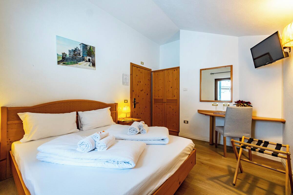 A hotel room with a wooden bed, a desk with a chair, a television mounted on the wall, and a painting on the wall.