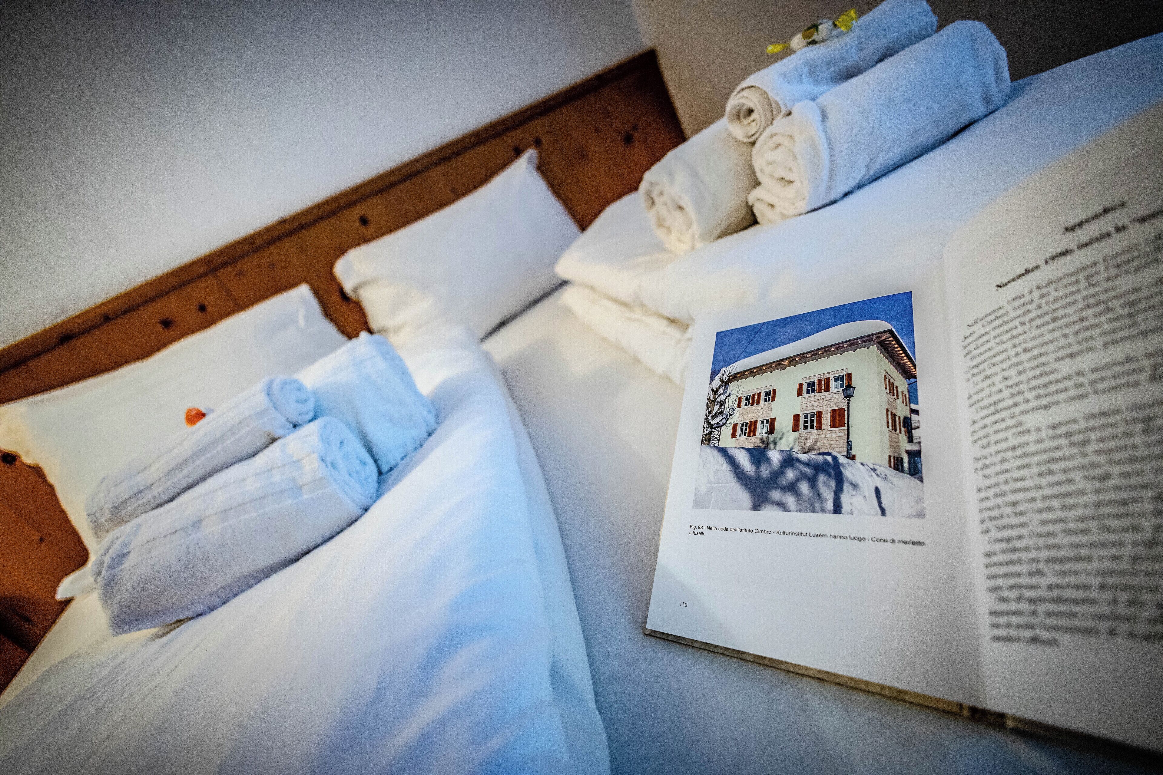 A neatly made bed with rolled towels and an open book on a wooden headboard.