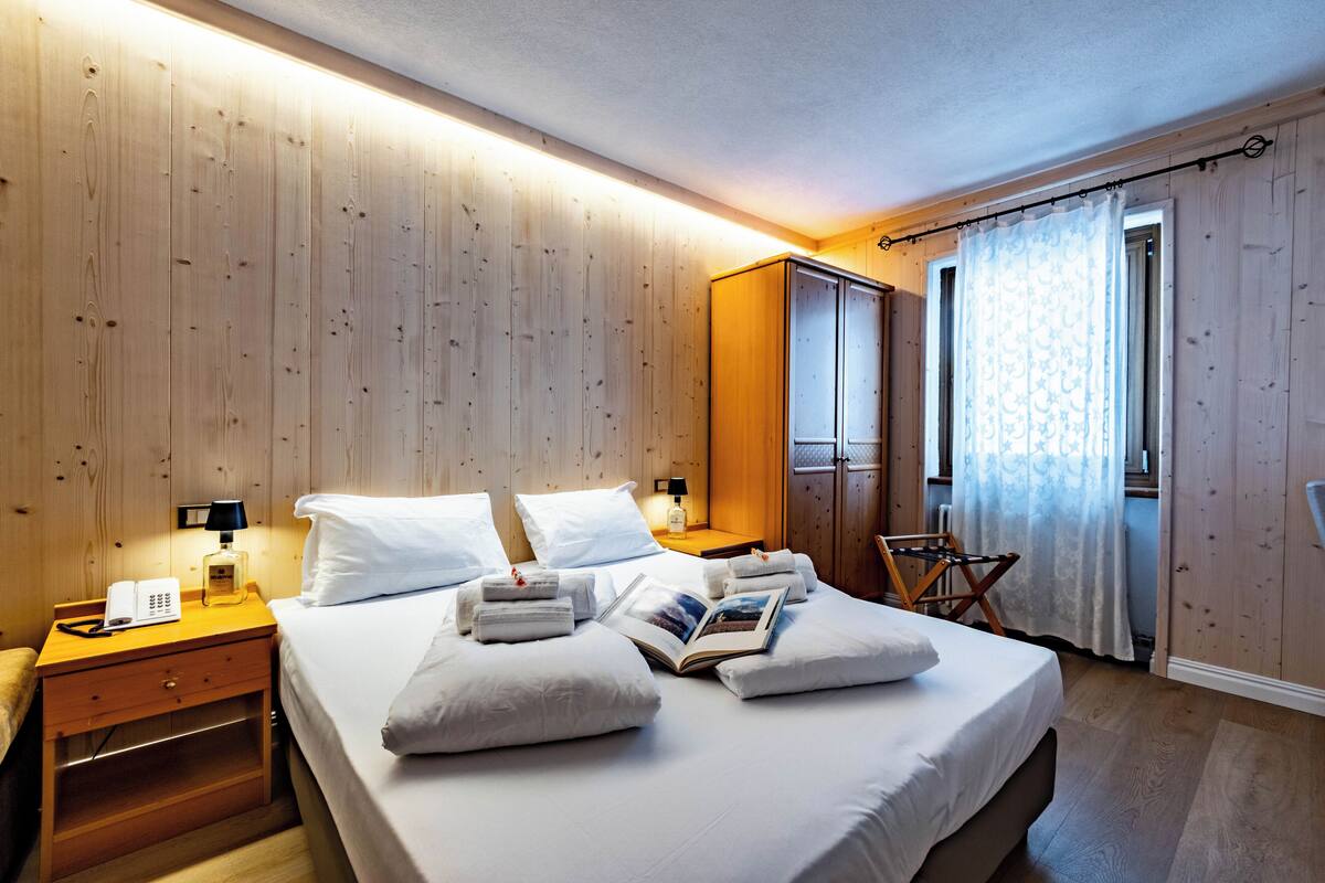 A hotel room with a wooden paneled wall, a bed with white linens, a nightstand with a lamp, a wardrobe, and a window with curtains.