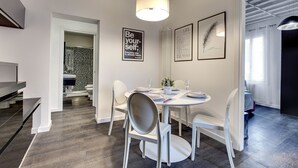 Un espace repas moderne avec une table ronde, des chaises blanches et un luminaire au plafond. Les murs sont ornés de citations encadrées, et on aperçoit une autre pièce avec une cabine de douche décorée.