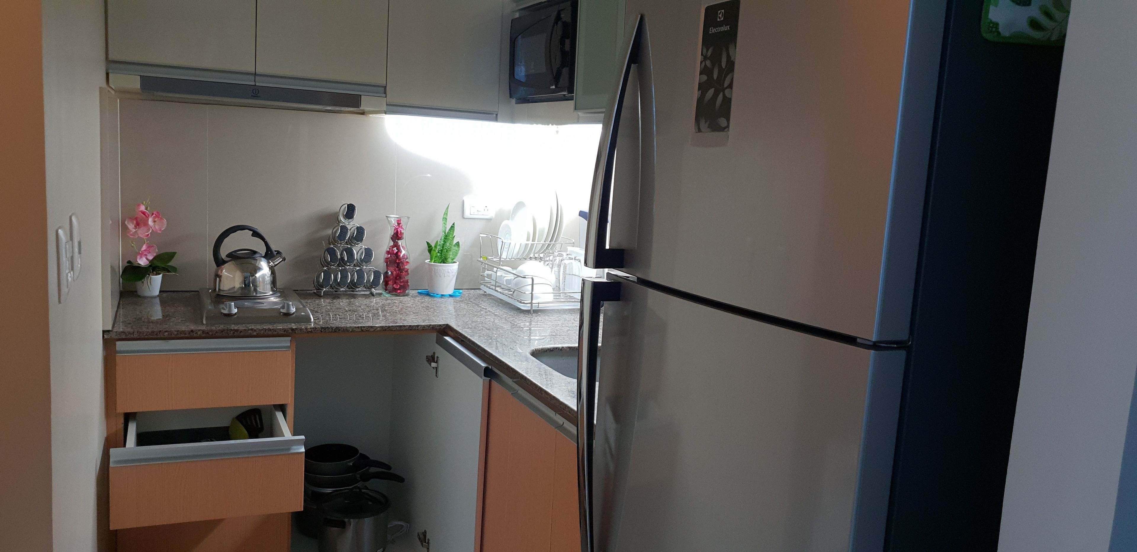 A compact kitchen with a refrigerator, microwave, and a small sink. There are cabinets, a kettle, and a potted plant on the counter.
