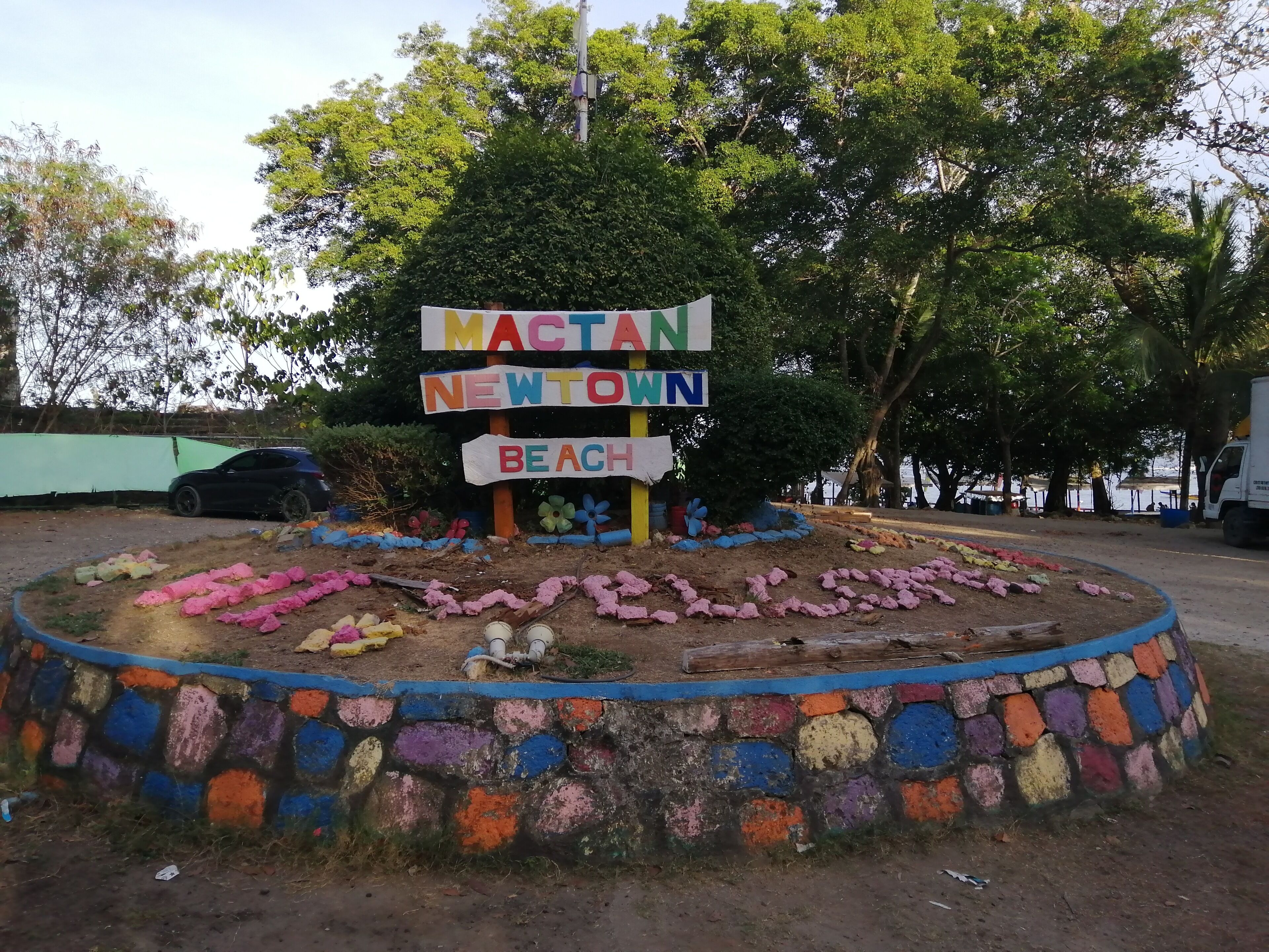 Une enseigne colorée sur laquelle on peut lire « MACTAN NEWTOWN BEACH », entourée d’un mur de pierres peintes.