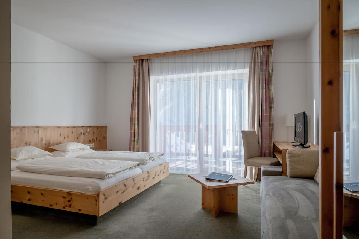 A hotel room with a wooden bed, a desk with a computer, a chair, and a large window with sheer curtains.