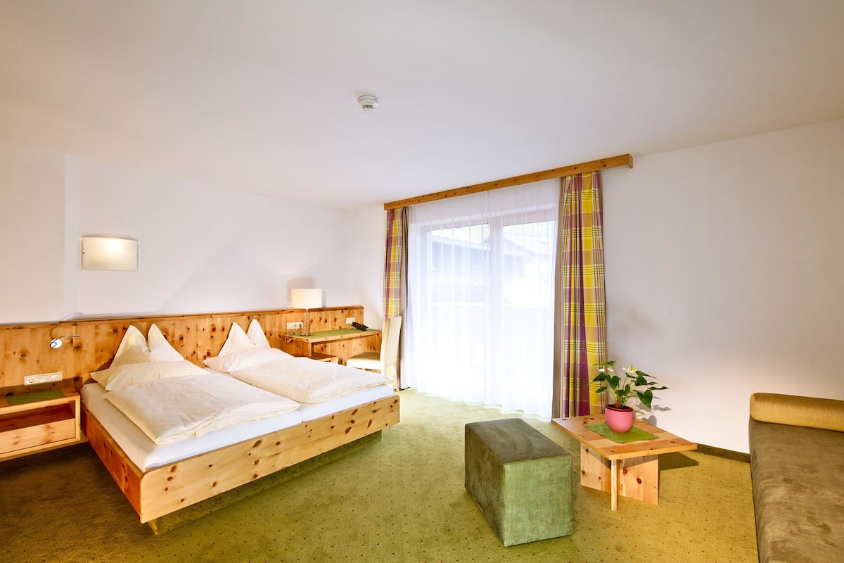 A hotel room with a wooden bed, a bedside table with a lamp, a small desk, a green ottoman, and a window with curtains.