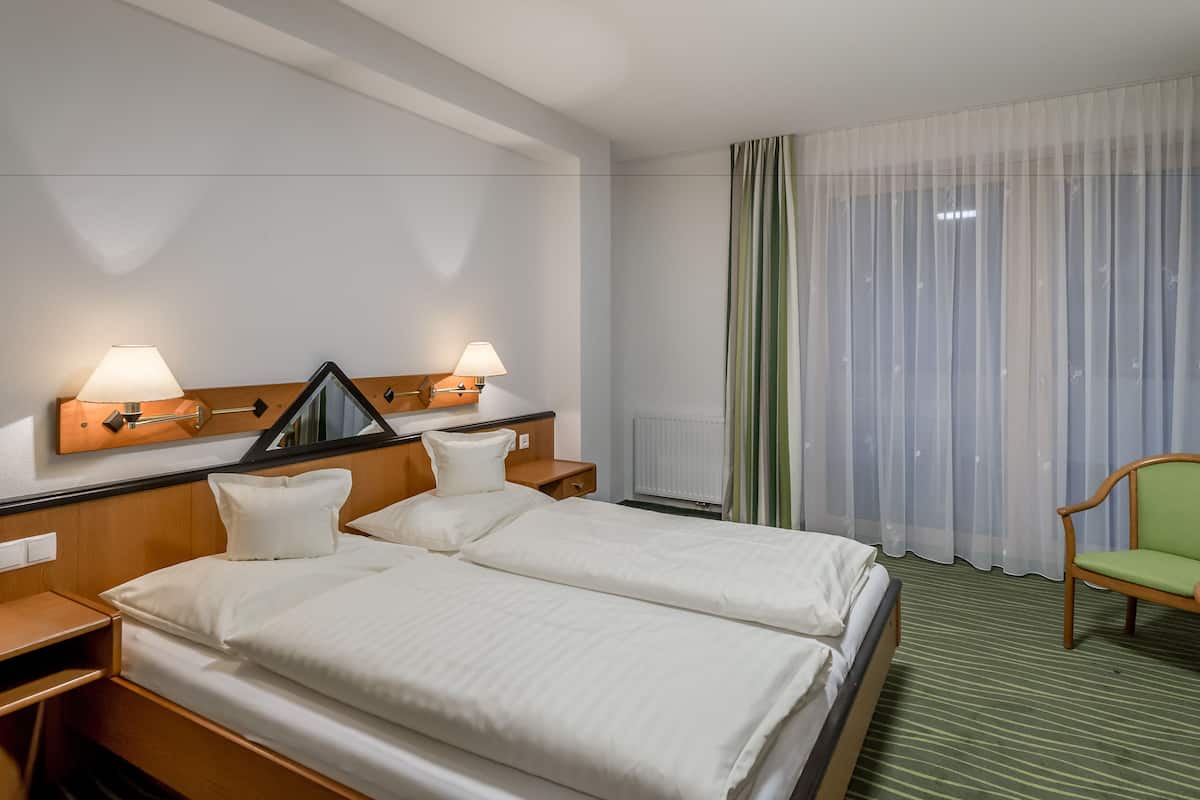 A hotel room with a double bed, two bedside tables, a mirror, a lamp, a radiator, a chair, and a window with sheer curtains.