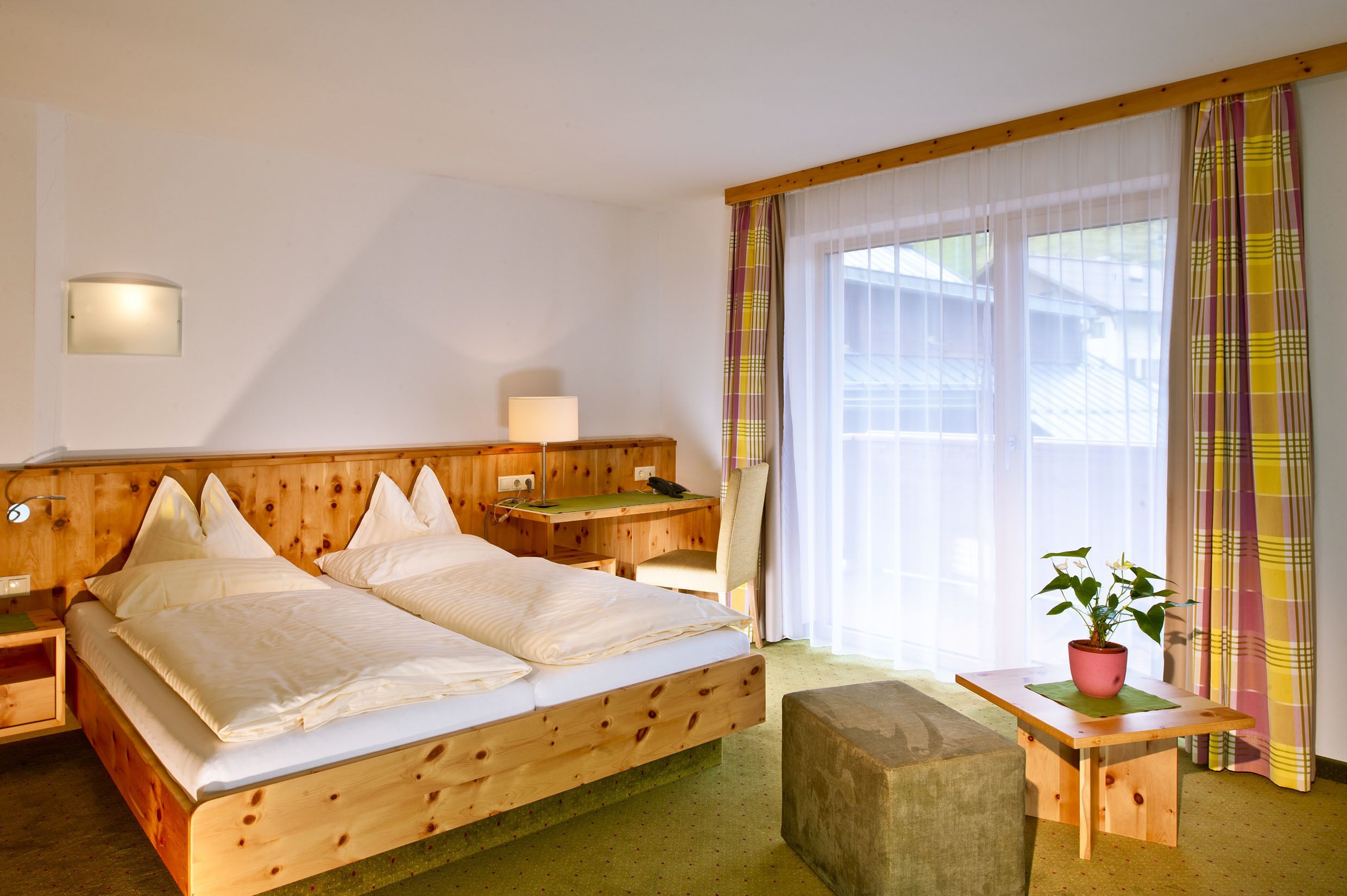 Ein hölzernes Bett mit Kissen, ein Nachttisch mit Lampe, ein kleiner Tisch mit Pflanze und ein Fenster mit Vorhängen.