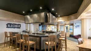 Un espace bar moderne avec un îlot central, des tabourets de bar et une enseigne indiquant « BLECHBAR ».