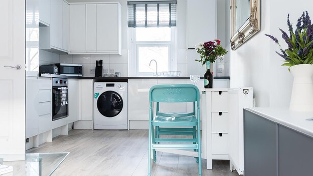 Une cuisine moderne avec des meubles blancs, un lave-linge et une chaise de couleur turquoise.